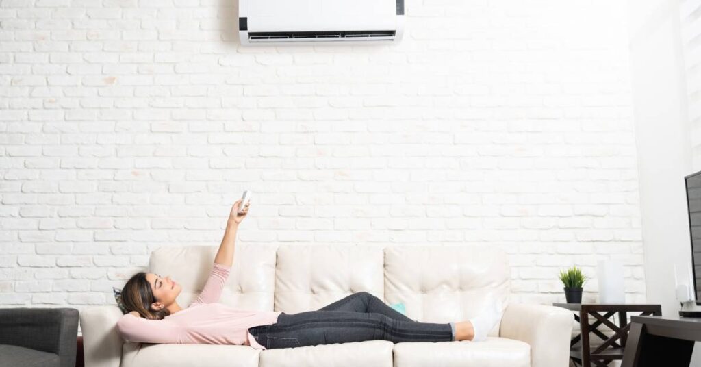 A woman lying on a cream-colored couch in a living room holds the remote to a mini split and points it toward the AC unit.