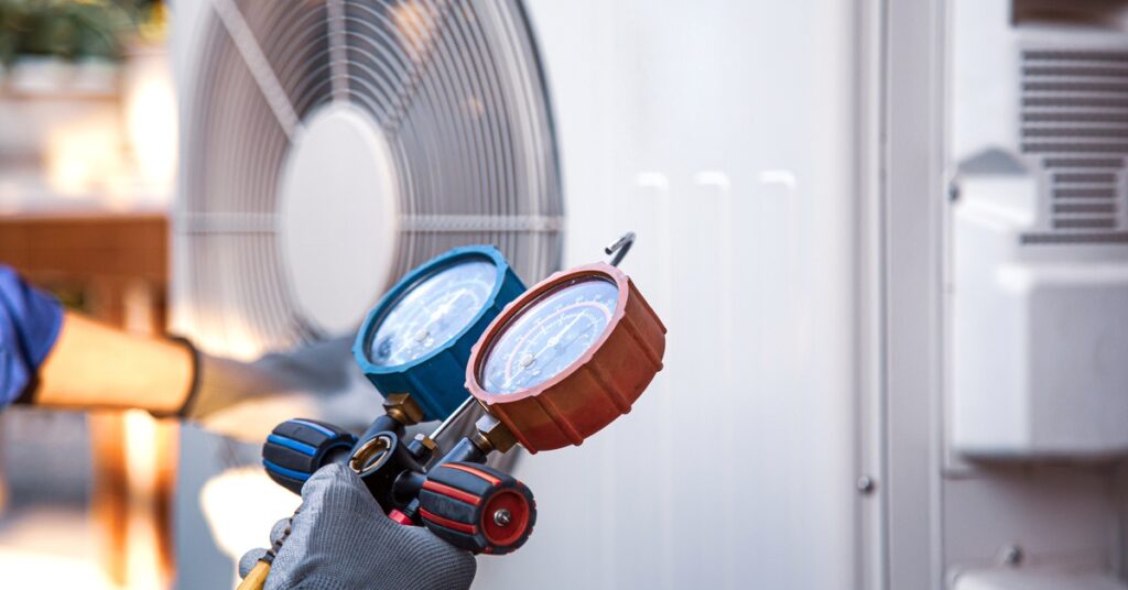 A gloved hand holding an HVAC pressure gauge. A large, slightly blurred, white HVAC system is in the background.