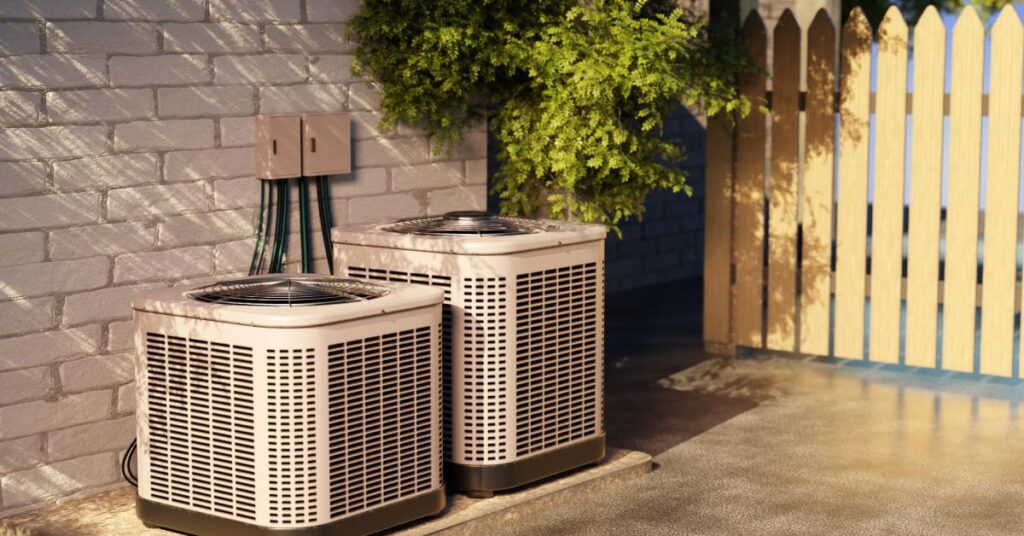 Two AC units sit beside one another on a concrete slab outside of a brick home; there’s a wood fence off to the side.