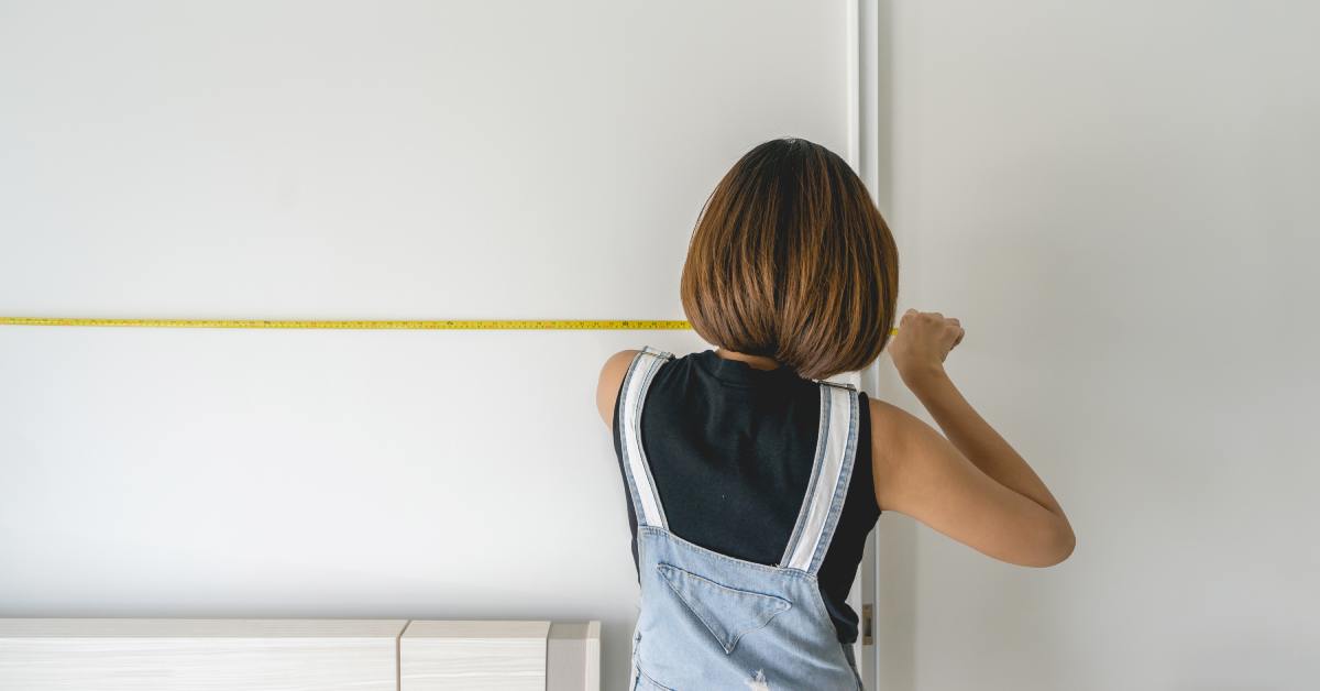 The backside of a woman in overalls and a black shirt. She’s holding an extended tape measure and facing a white wall.