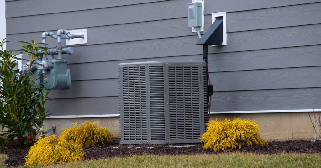 An outdoor air conditioning unit sits in mulch beside a gray-sided home, with small plants growing next to the system.