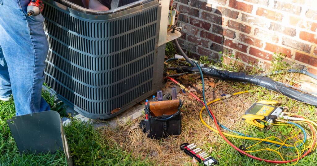 An unseen person reaches into an outdoor AC unit with the top removed; cables and a tool bag rest on the ground nearby.