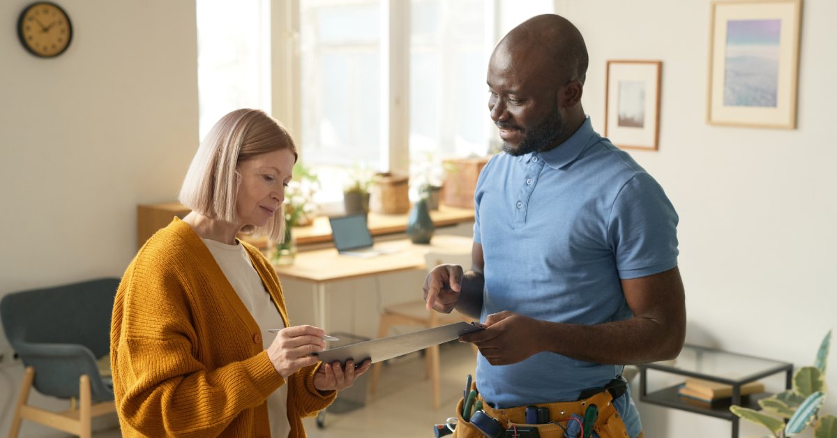 A man in a tool belt and a woman with a clipboard converse in a room furnished with a desk, a chair, and a side table.