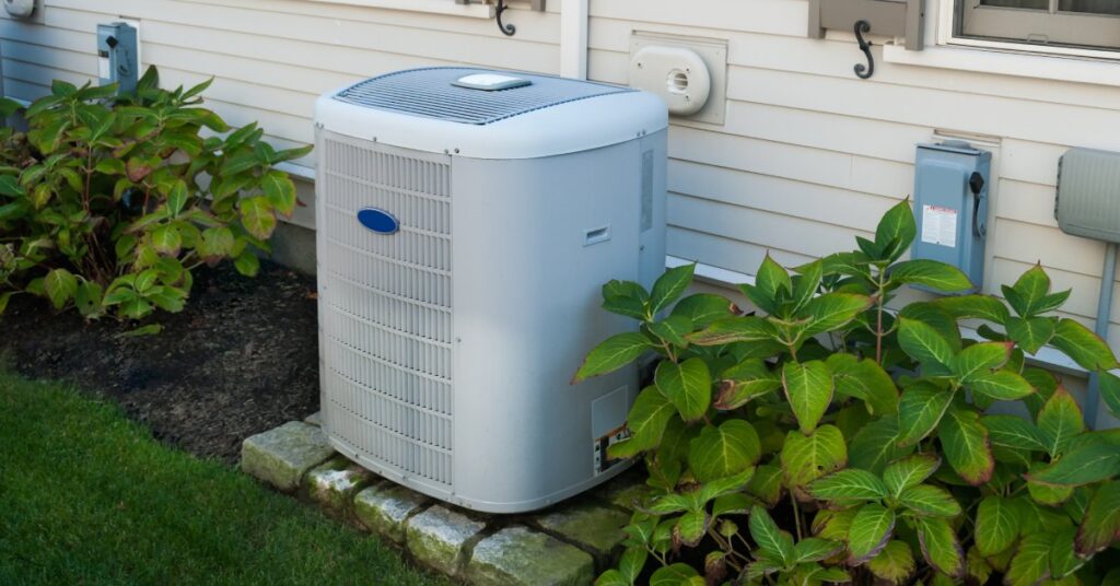 An outdoor air conditioning unit sits on gray bricks beside a home with cream-colored siding and small bushes.