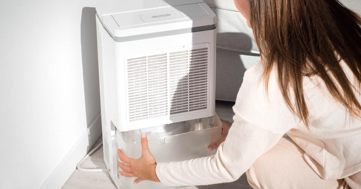 A woman kneels down and holds the water container in the slot of a white dehumidifier; a plain wall and floor are visible.