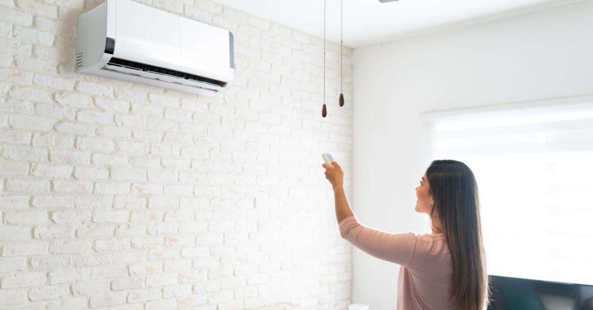 A woman adjusts a white air conditioner on a light wall with a remote; a sofa and ceiling fan are in the background.