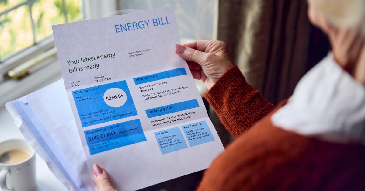 A partially seen person in an orange sweater holds an energy bill, showing $366.85 due, with a blurred indoor background.