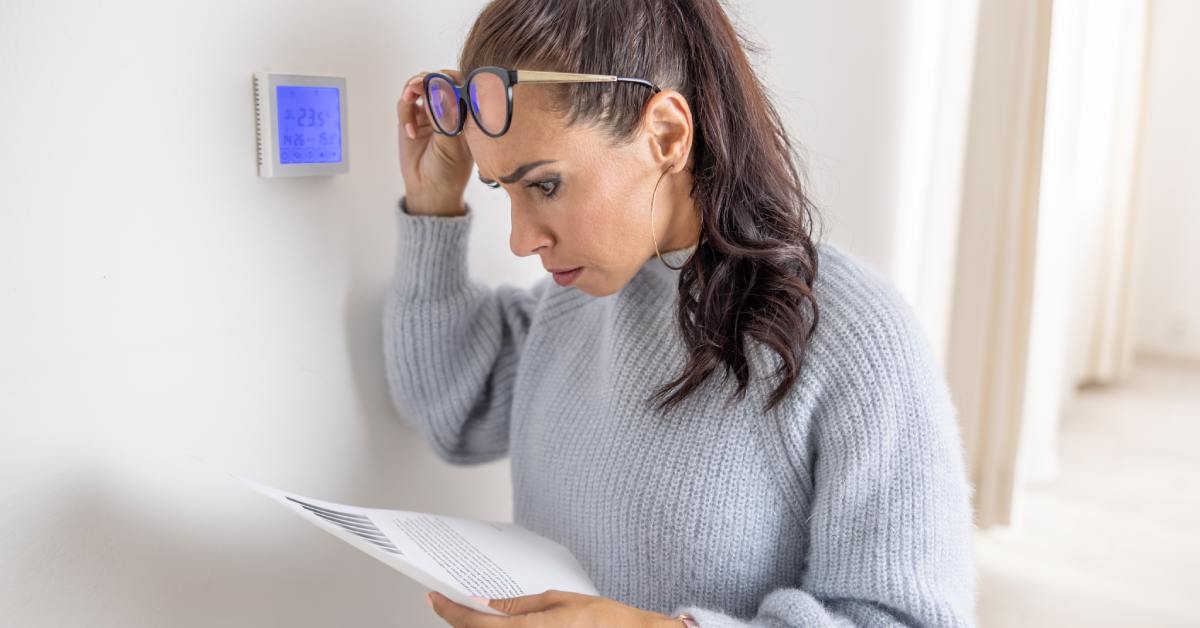 A woman in glasses and a gray sweater looks at a bill with wide eyes and stands near a thermostat reading 23.9°C.