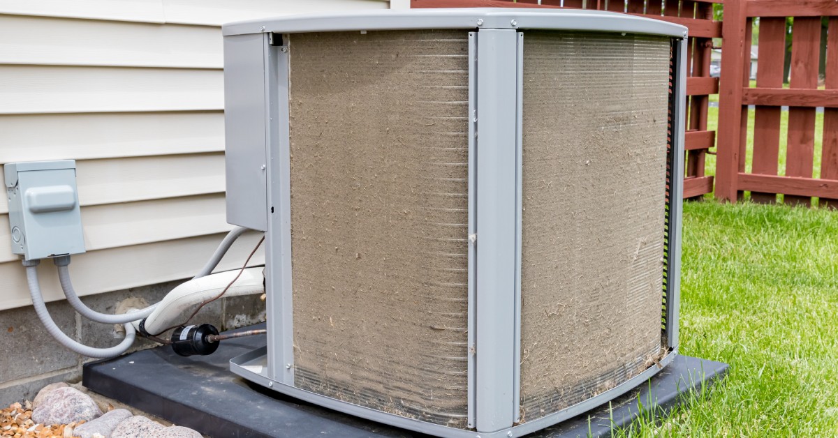 A dirty outdoor air conditioning unit with condenser coils clogged by dirt and grass debris, placed near a house wall.