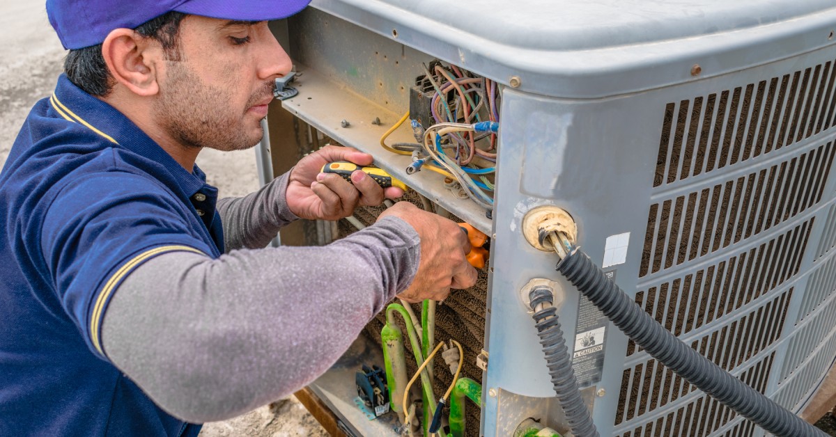 An electrician in a blue uniform and cap repairs an air conditioning unit, using tools to fix internal components.