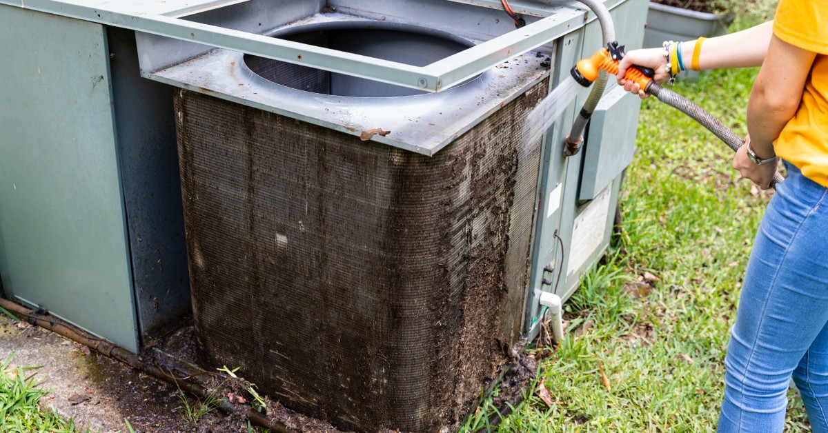 A person in a yellow shirt and jeans cleans dirty condenser coils on an outdoor air conditioning unit using a garden hose.