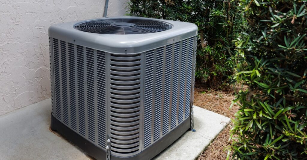 A modern HVAC unit on a concrete slab outside a house, surrounded by green shrubs.