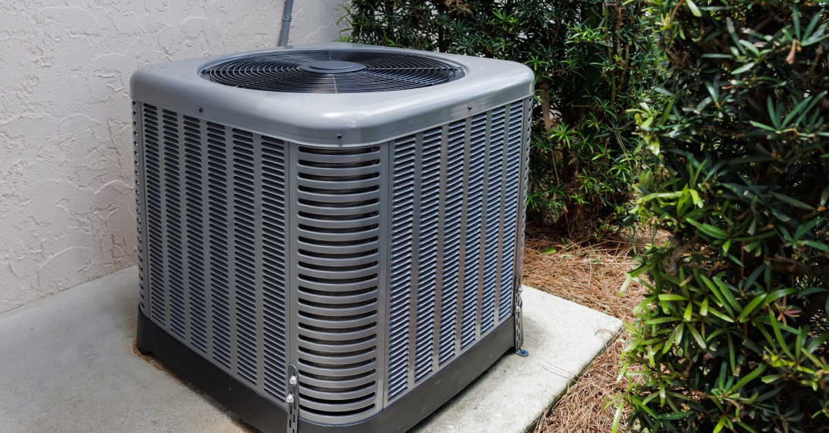 A modern HVAC unit on a concrete slab outside a house, surrounded by green shrubs.