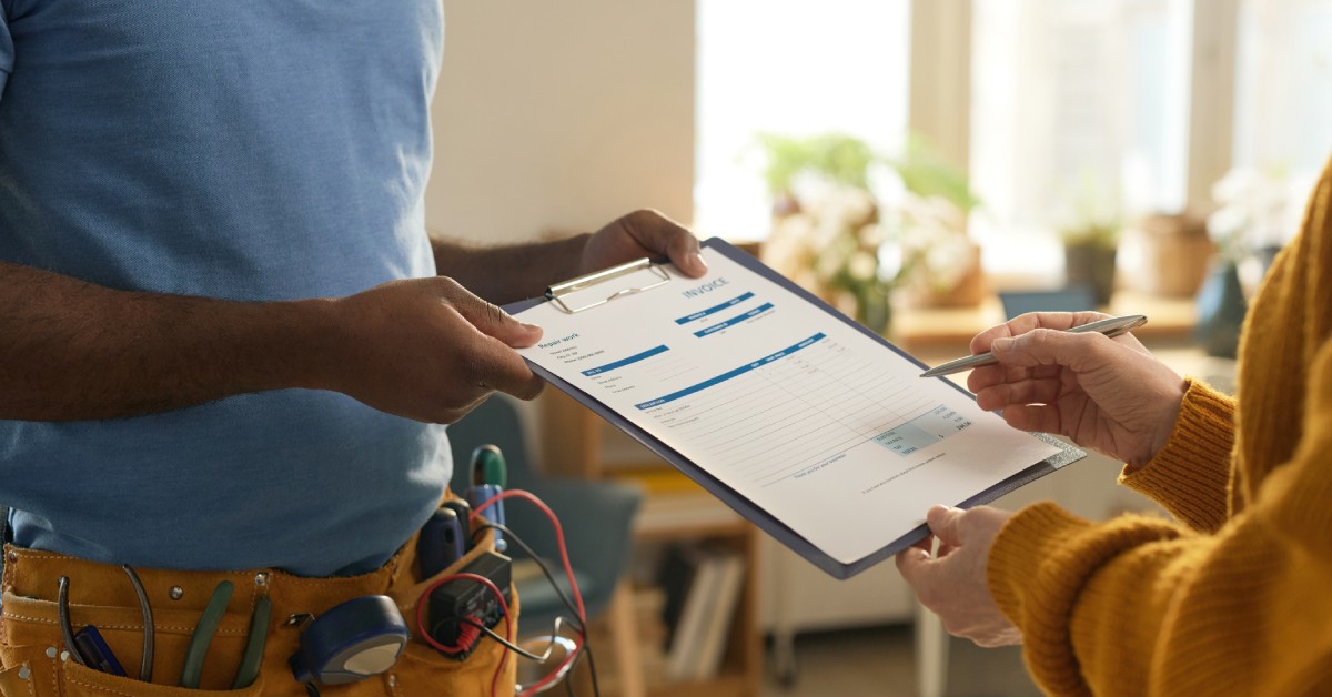 A close-up side view of a man with a toolbelt handing a clipboard to a client signing a contract for home repairs.