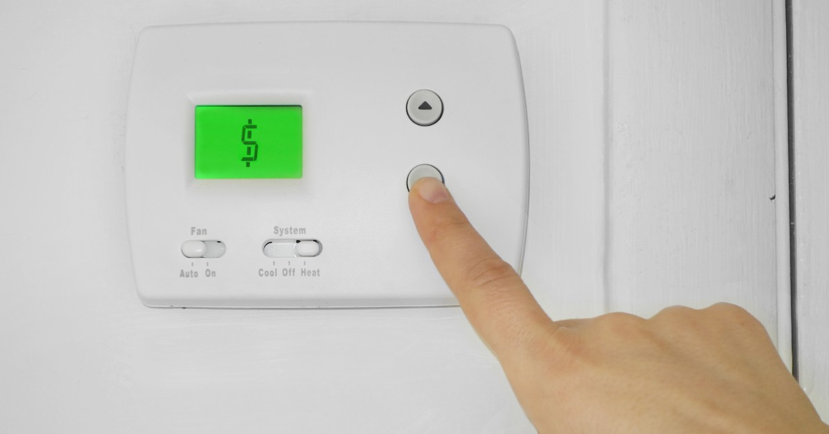 A white digital thermostat with a green display showing a dollar sign, with a hand pressing one of its buttons.